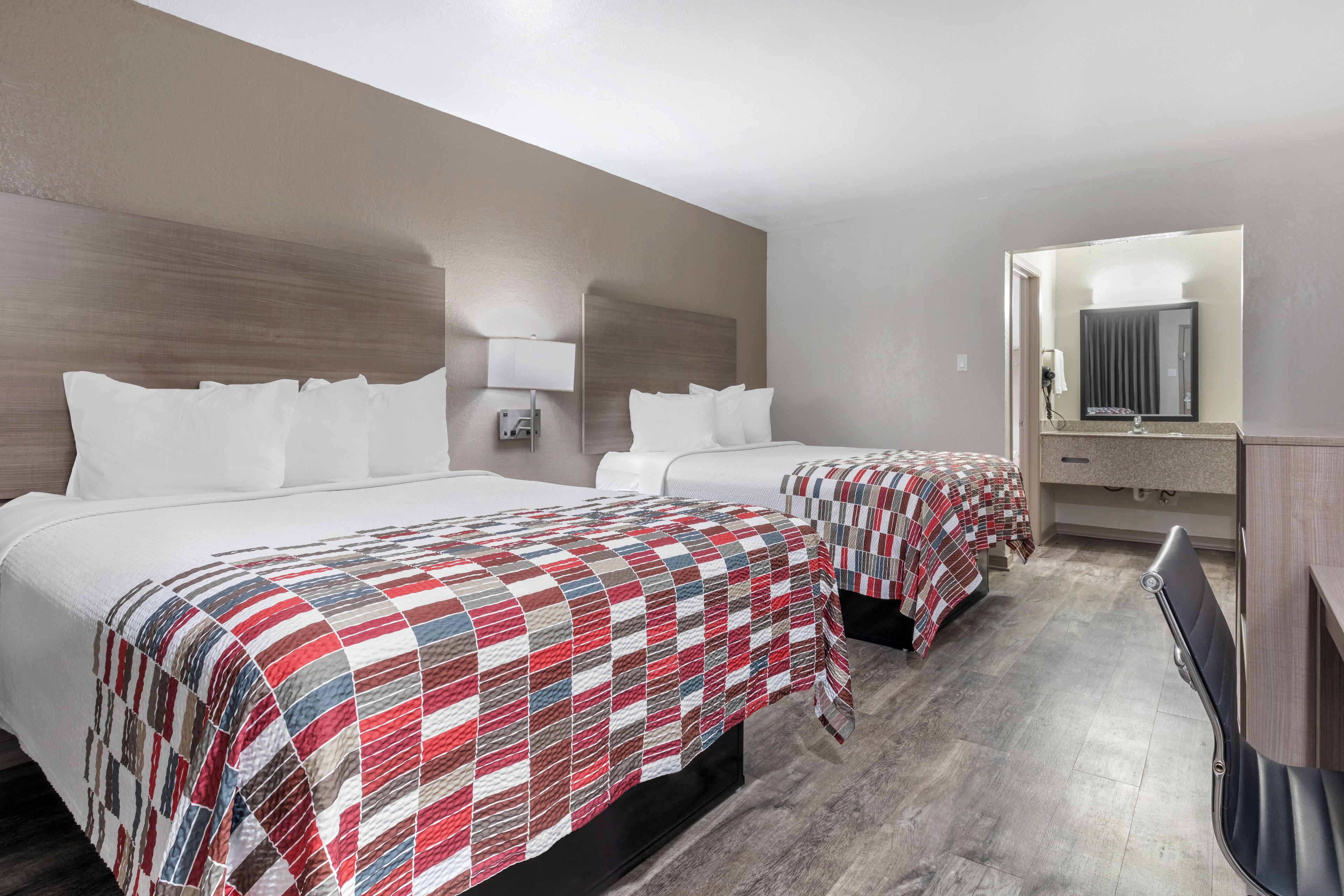 A hotel room with two beds, a bedside table with a lamp, a mirror, and a bathroom visible through an open door.