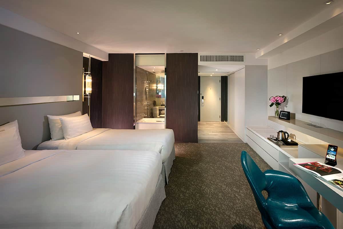 Quarto de hotel moderno com duas camas, uma escrivaninha e uma televisão de tela plana. Um banheiro é visível através de uma porta aberta.