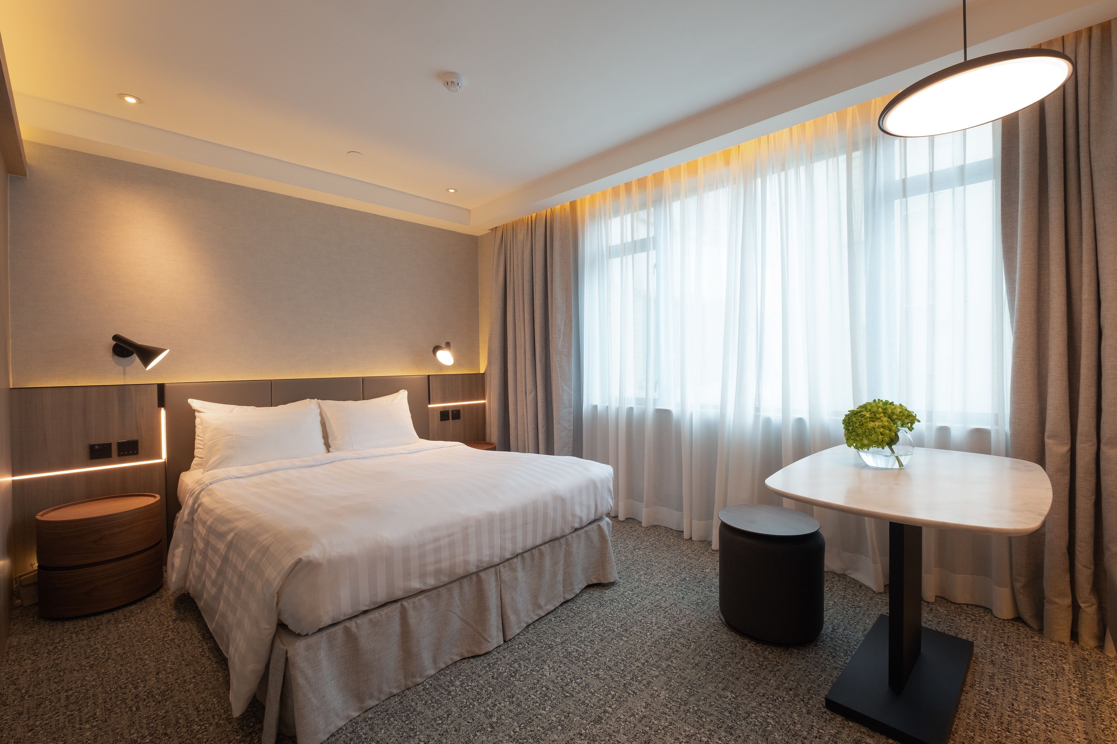A modern hotel room with a large bed, bedside tables, a round table with a vase, and a view of the outdoors through sheer curtains.