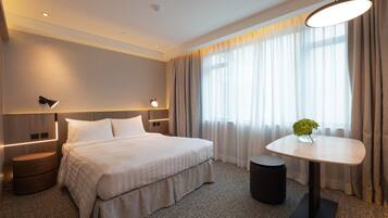 A modern hotel room with a large bed, bedside tables, a round table with a vase, and a view of the outdoors through sheer curtains.