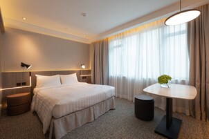A modern hotel room with a large bed, bedside tables, a round table with a vase, and a view of the outdoors through sheer curtains.