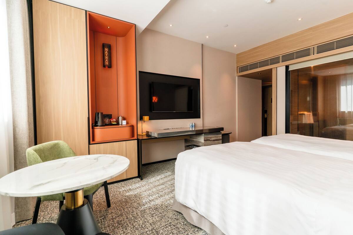 Quarto de hotel moderno com uma cama grande, uma mesa de trabalho com televisão de tela plana, uma mesinha redonda e uma cadeira verde.