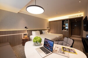 A modern hotel room with a bed, a desk with a laptop, a magazine, and a small plant.