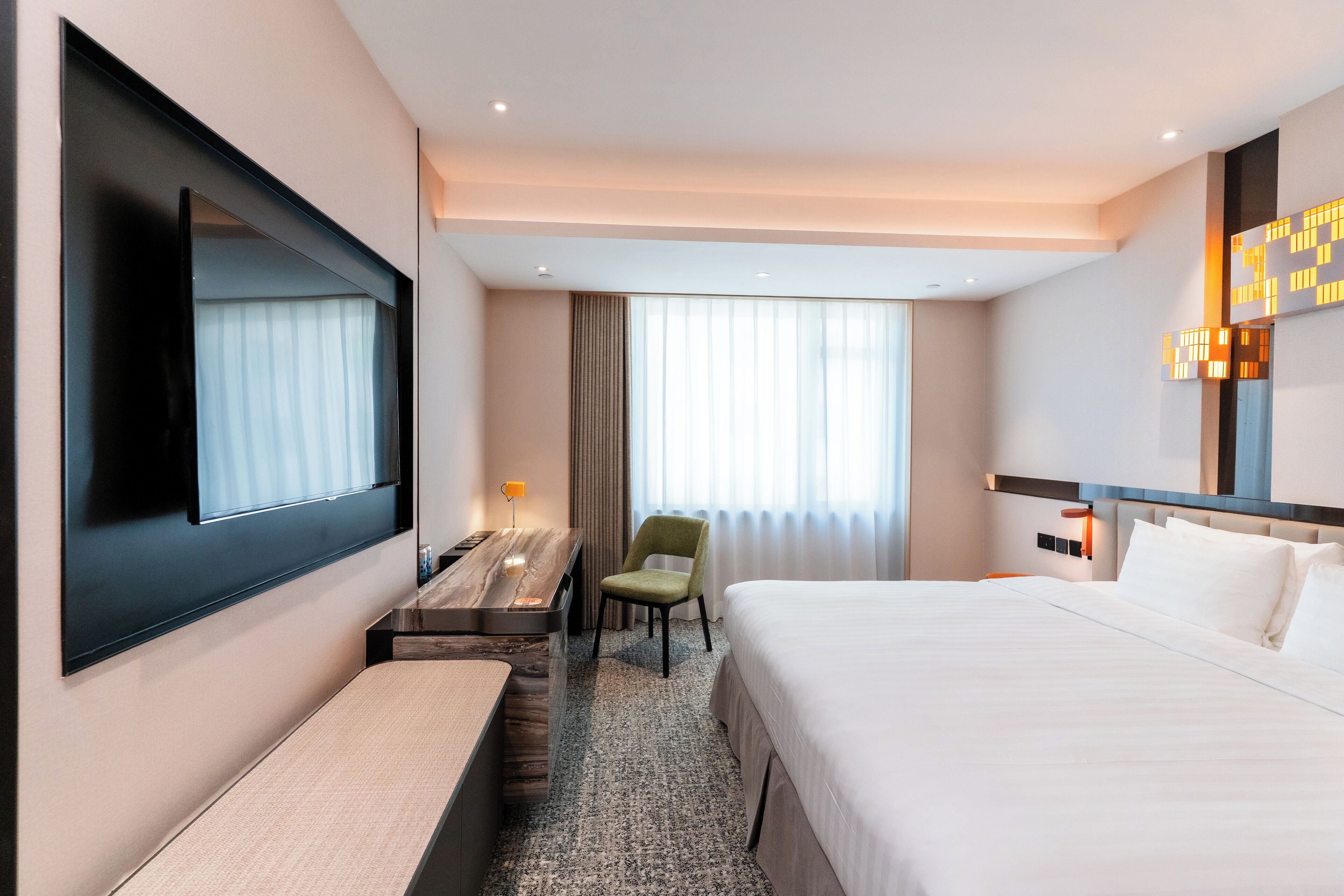 A modern hotel room with a large bed, a desk, a chair, and a TV mounted on the wall.