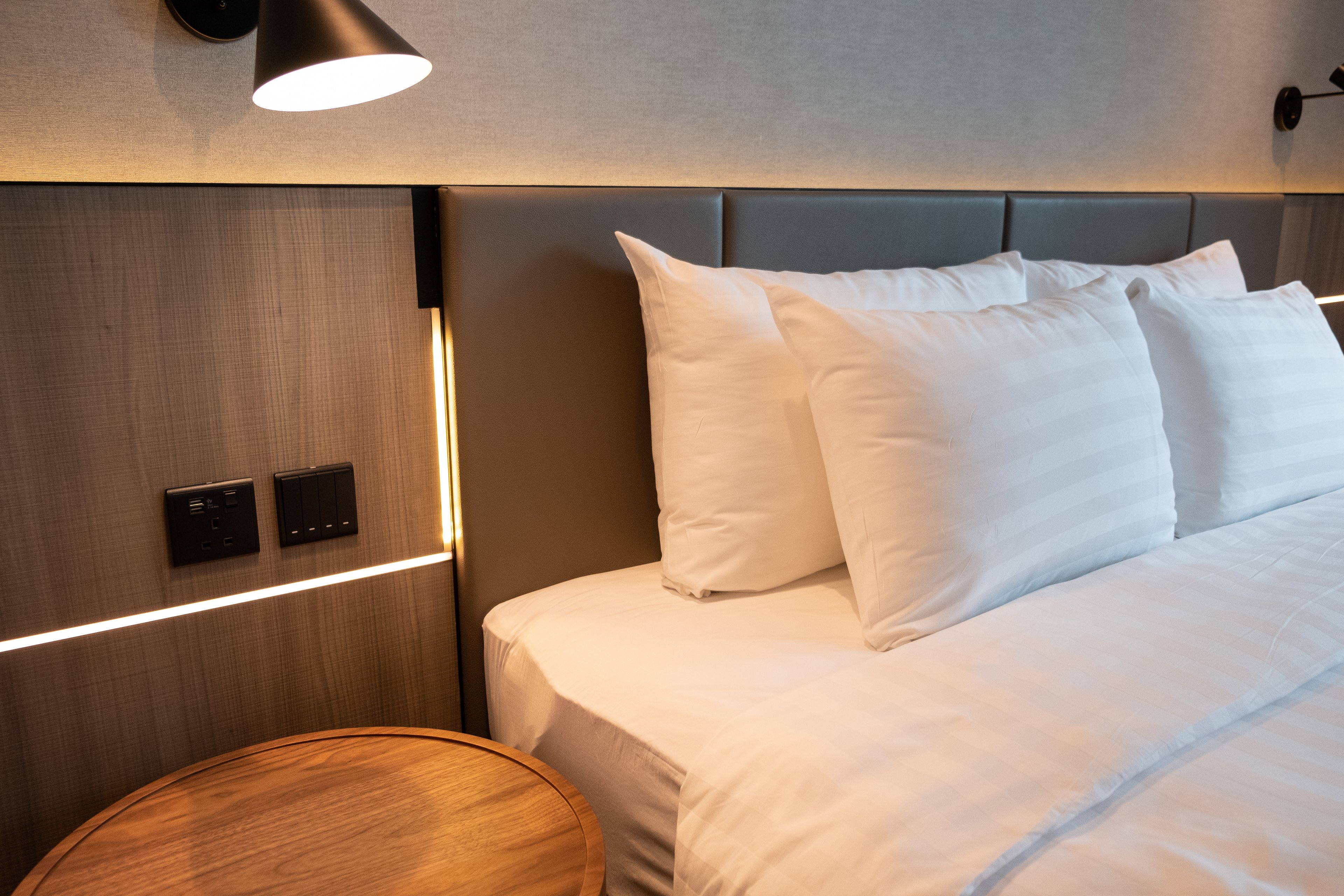 Una cama bien tendida con sábanas blancas y un cabecero, una mesita de madera redonda y una lámpara de pared.