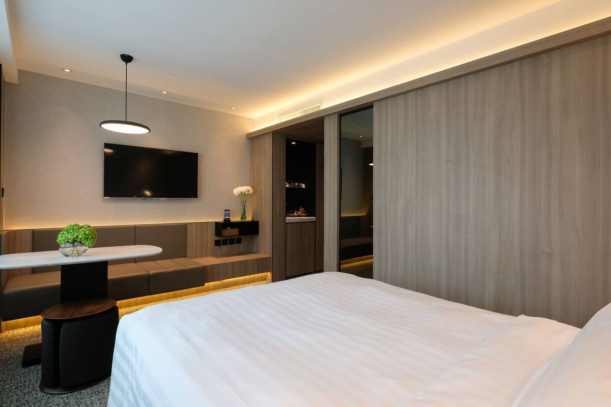 Quarto de hotel moderno com uma cama grande, televisão de tela plana na parede, área de estar com mesa e cadeiras, e porta deslizante para uma varanda.