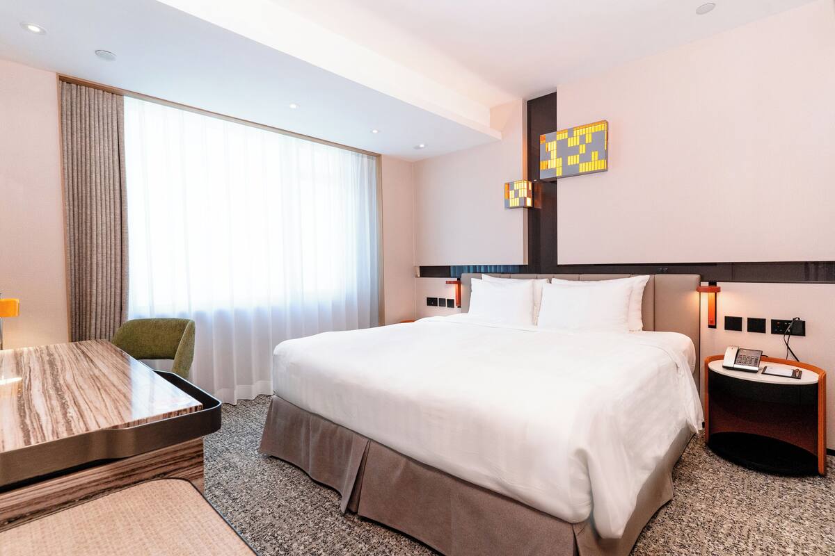 Quarto de hotel com cama grande, escrivaninha, cadeira, mesa de cabeceira com telefone e luminária na parede.