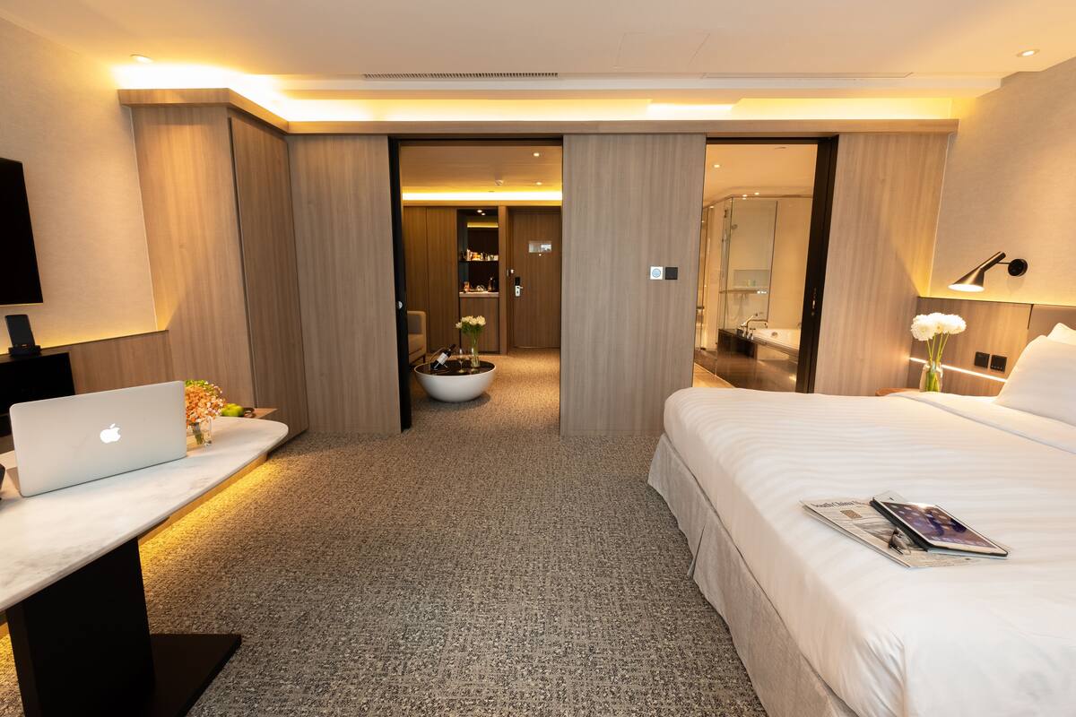 Quarto de hotel moderno com cama, escrivaninha com laptop, mesa de cabeceira com vaso e banheiro visível através de uma porta aberta.