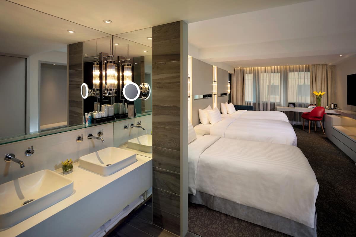 Quarto de hotel moderno com um espelho grande, duas pias brancas, toucador, cama com roupa de cama branca, secretária com cadeira e televisão.