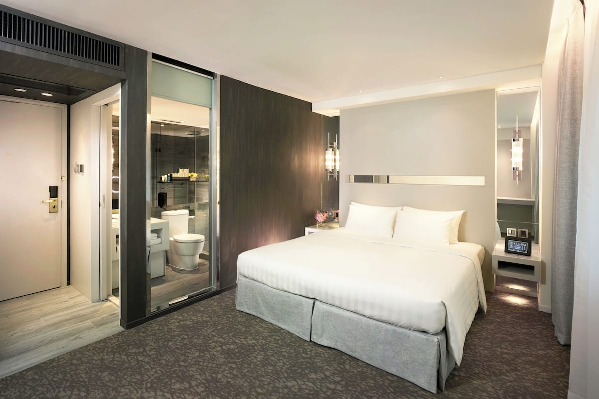 Quarto de hotel moderno com uma cama grande, um banheiro com paredes de vidro e uma mesa de cabeceira com relógio digital.