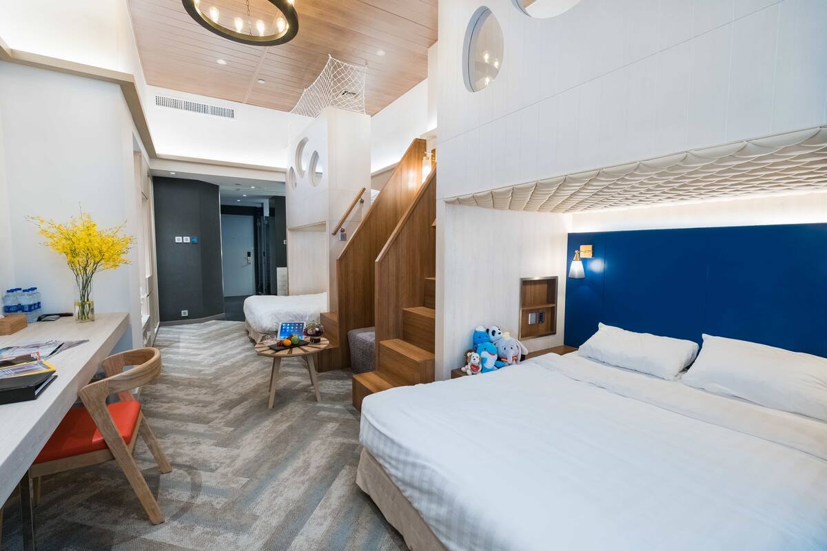 Quarto de hotel moderno com uma cama grande, uma escrivaninha, uma cadeira, uma mesinha com um vaso de flores e uma escada.