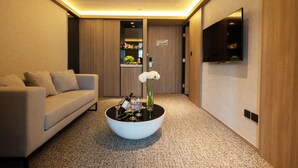 A modern living room with a sofa, a coffee table with a vase of flowers, a flat-screen TV mounted on the wall, and a wooden door leading to another room. - Nathan Hotel (Kowloon)