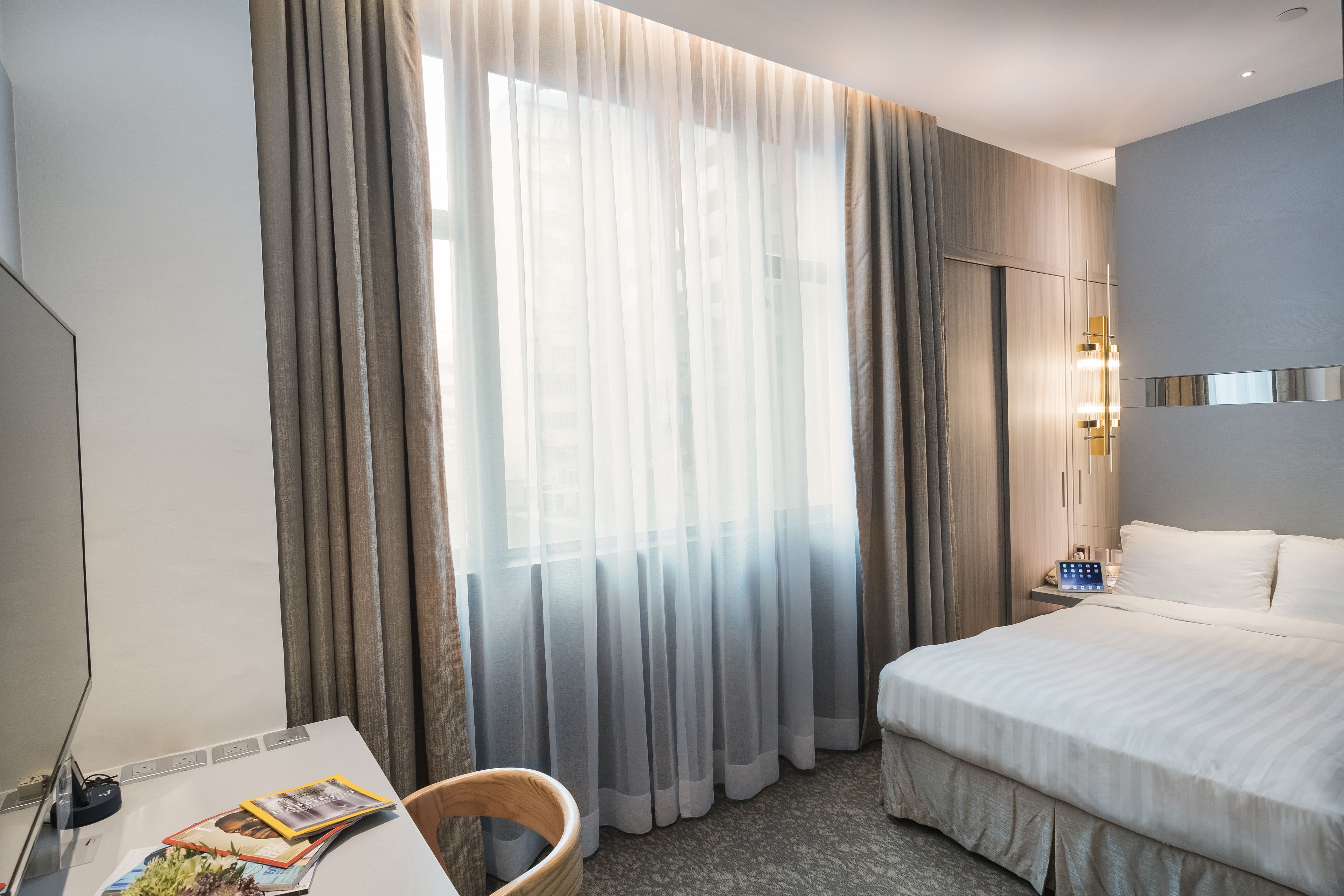 A hotel room with a large bed, a desk with a computer, and a window with sheer curtains.
