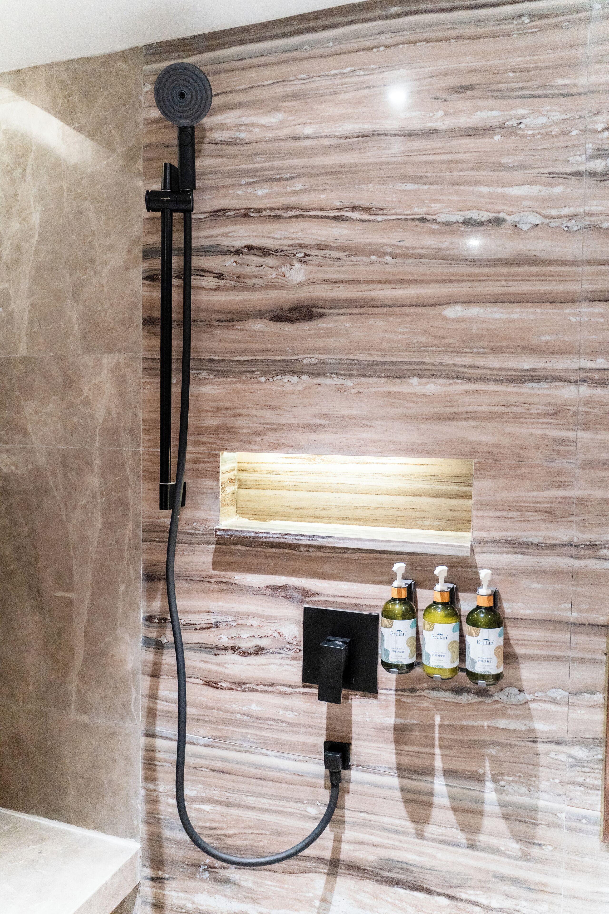 A bathroom with a marble wall, a handheld showerhead, and three bottles of Erulan on a shelf.