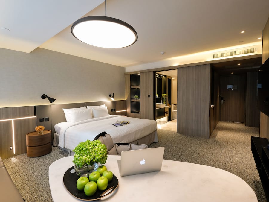 Una habitación de hotel moderna con una cama, una mesita de noche con una computadora portátil, un cuenco de manzanas y un jarrón de flores.