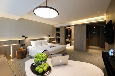 A modern hotel room with a bed, bedside table with a laptop, a bowl of apples, and a vase of flowers.