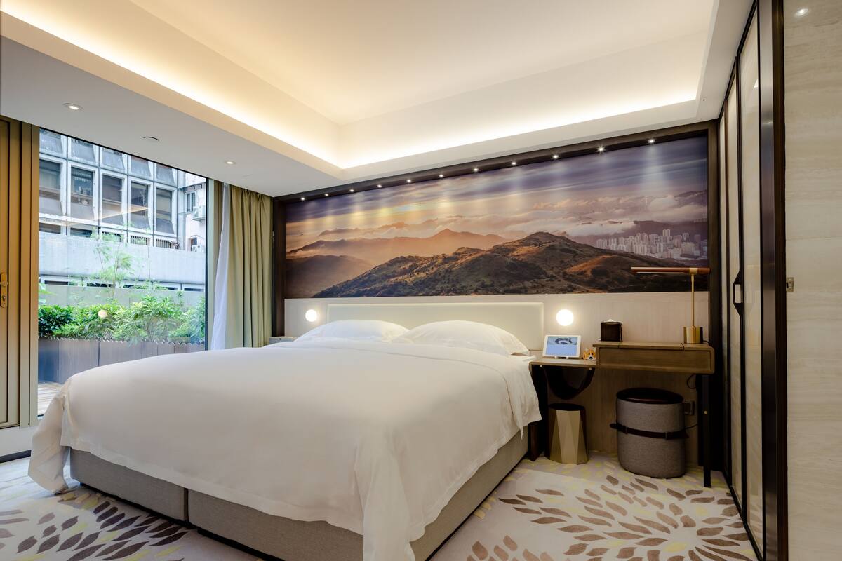 A modern hotel room with a large bed, a desk with a computer, and a scenic mountain view on the wall.