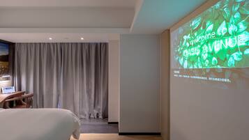 A hotel room with a bed, a desk, a TV, and a projection screen displaying a welcome message.