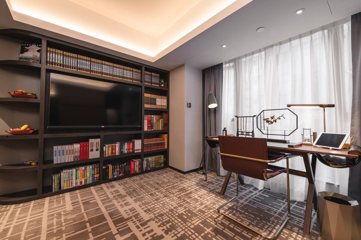 A modern hotel room with a large flat-screen TV, a bookshelf filled with books, a desk with a laptop, and a patterned carpet.