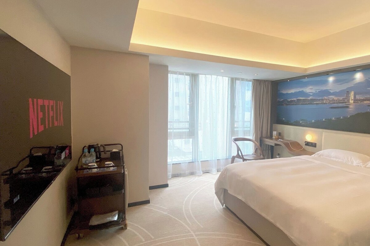 A hotel room with a bed, a desk, a chair, a minibar, and a large window with a cityscape view.