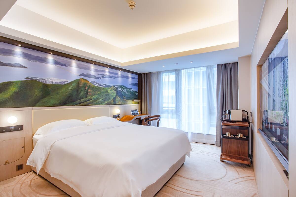 A hotel room with a large bed, a desk with a chair, a nightstand, a wall-mounted TV, and a window with curtains.