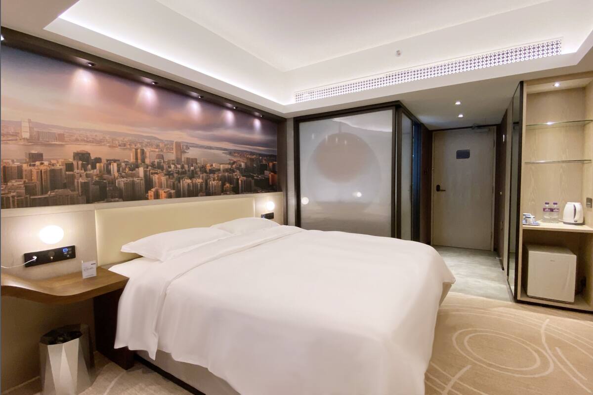 A hotel room with a large bed, a cityscape mural on the wall, a desk with a lamp, and a minibar.