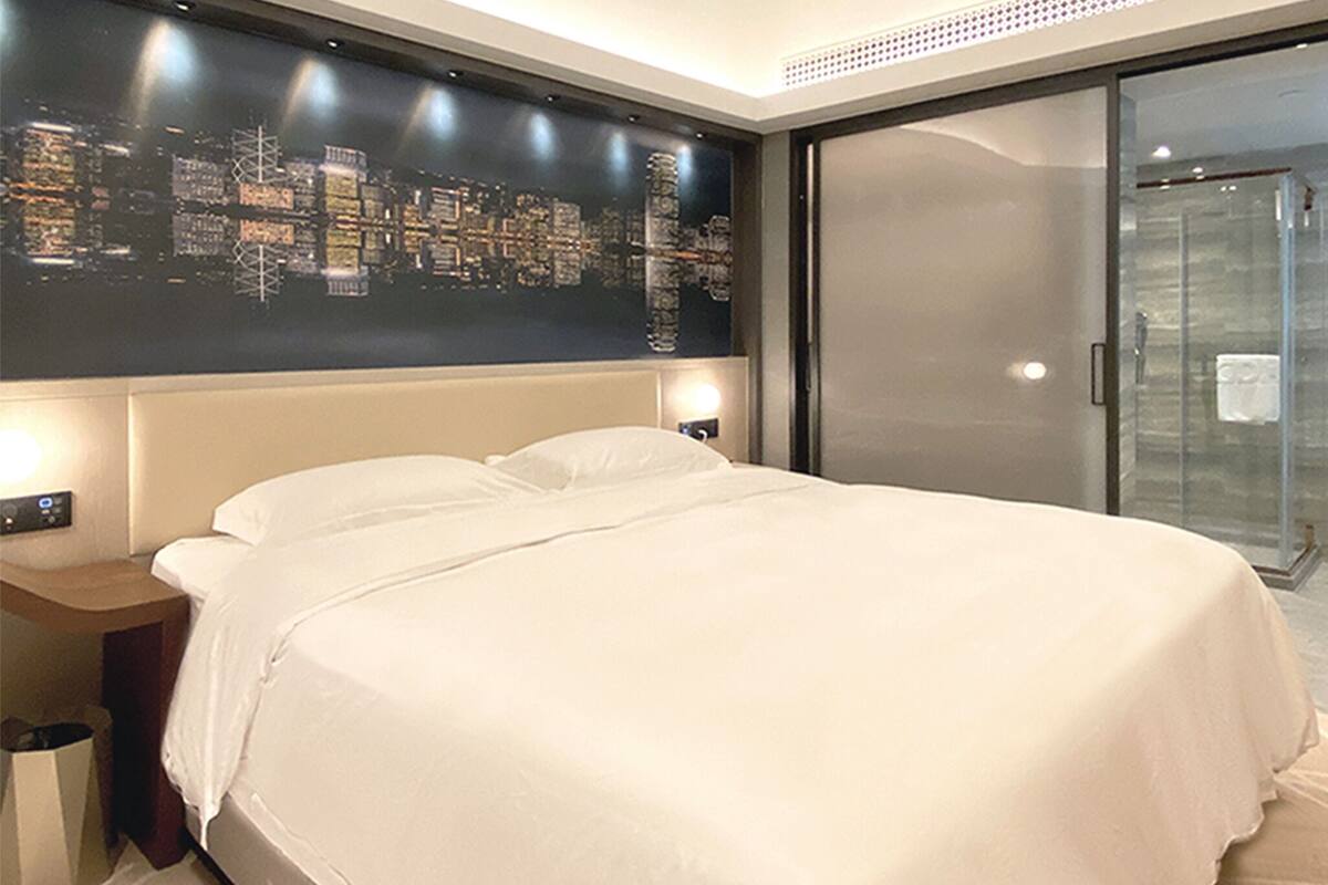 A hotel room with a large bed, a cityscape artwork on the wall, a glass shower door, and a nightstand with a lamp.