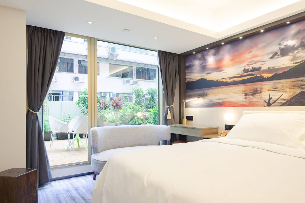 A hotel room with a bed, a desk, a chair, and a view of a balcony with plants and a building in the background.