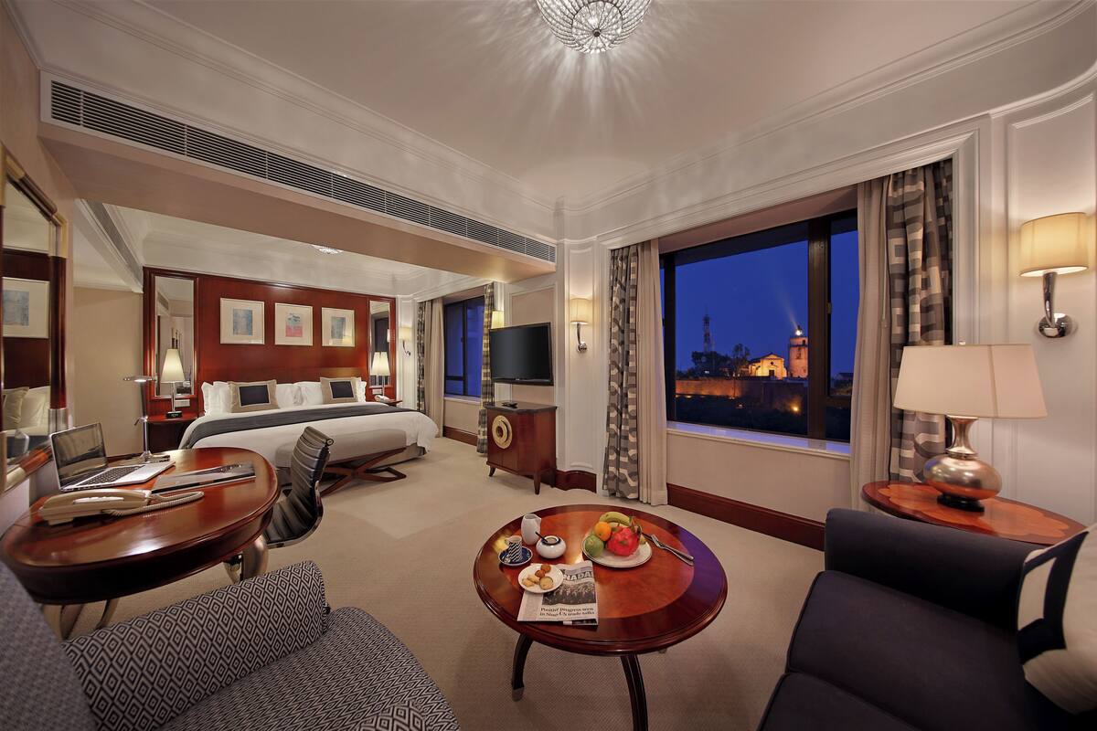 A hotel room with a bed, a desk with a laptop, a dining table with fruit, and a large window with a city view.