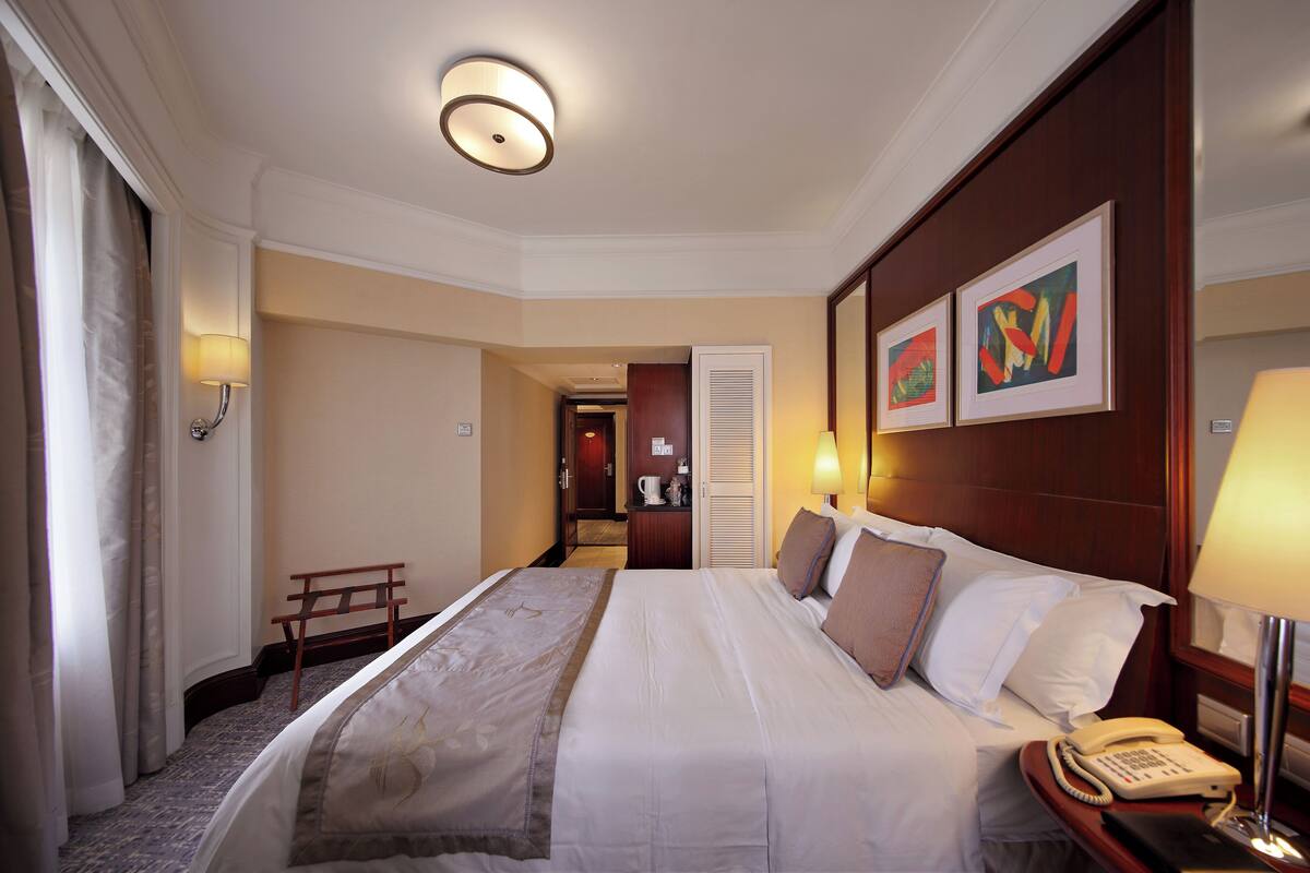 A hotel room with a neatly made bed, bedside table with a lamp, a telephone, and framed artwork on the wall.