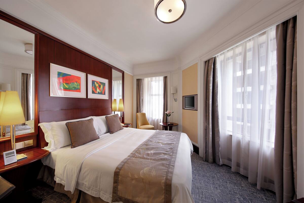 A hotel room with a large bed, a desk with a lamp, a chair, a TV, and a window with curtains.