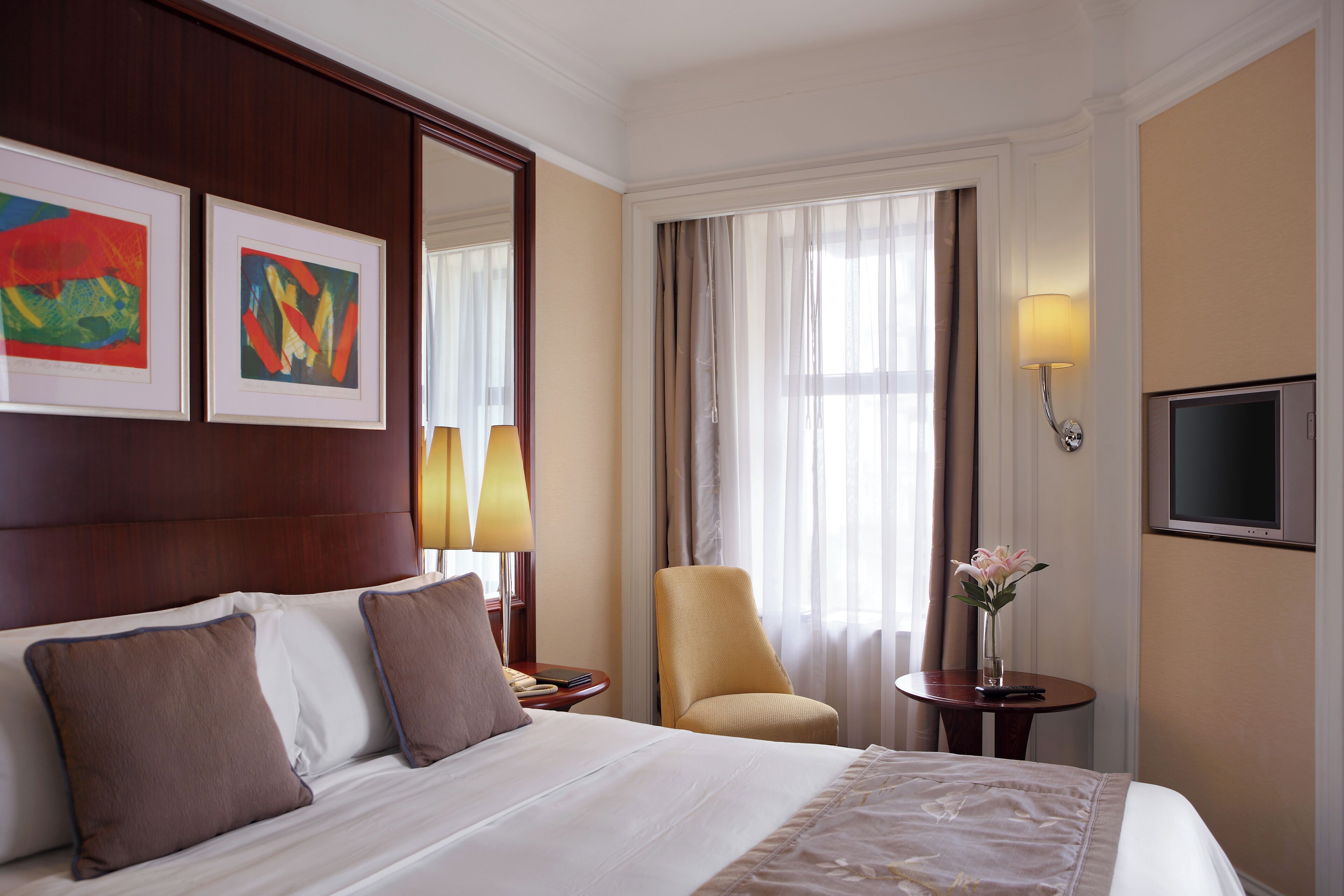 A hotel room with a large bed, two pillows, a nightstand, a chair, a table with a vase of flowers, a TV, and two framed artworks on the wall.