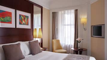 Ein Hotelzimmer mit einem großen Bett, zwei Kissen, einem Nachttisch, einem Sessel, einem Tisch mit Blumenvase, einem Fernseher und zwei gerahmten Kunstwerken an der Wand.