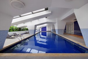 Una piscina interior con agua azul cristalina, un techo iluminado por el sol y vistas a la vegetación exterior.