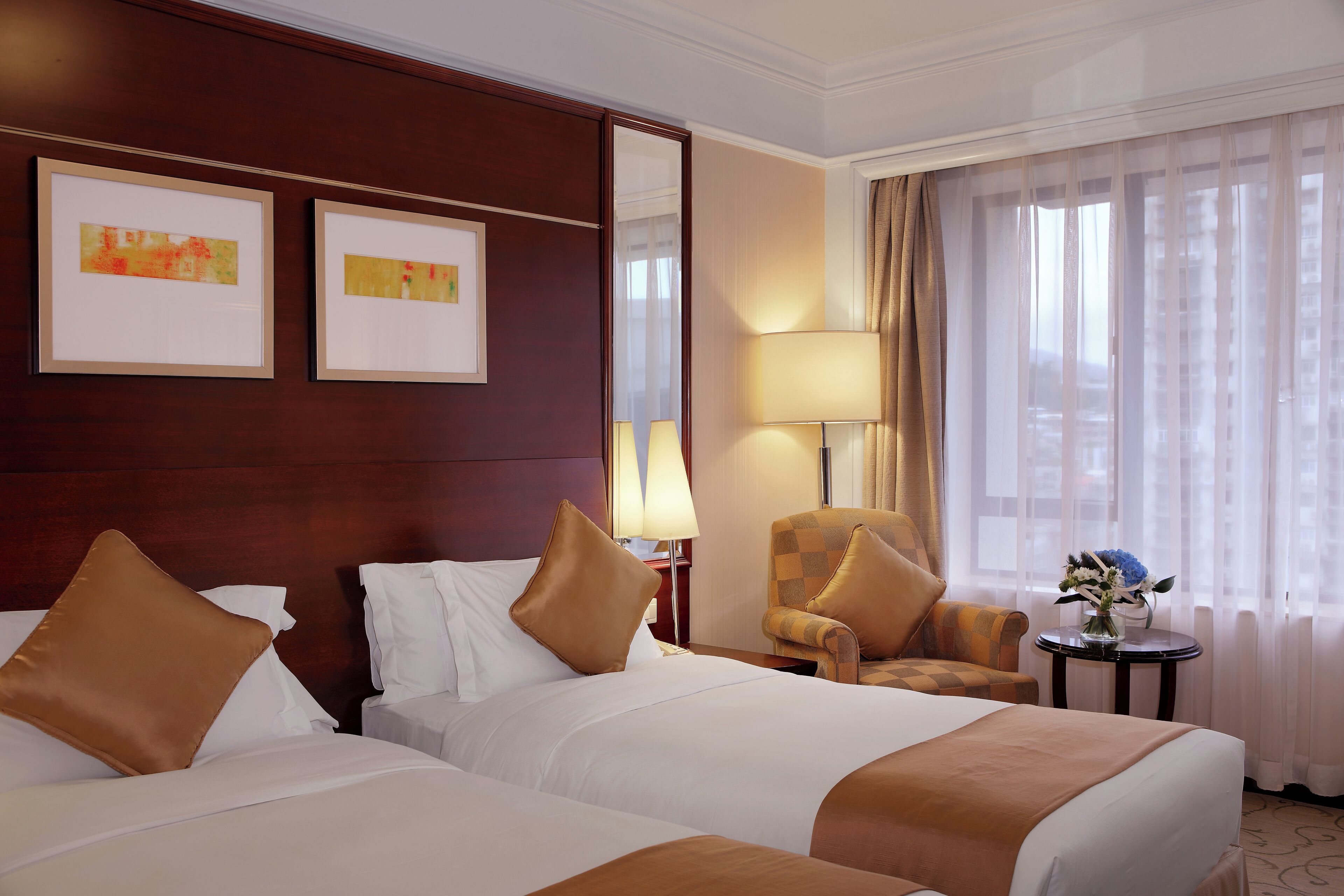 A hotel room with two beds, a sitting area with a chair, a lamp, and a small table with a vase of flowers.