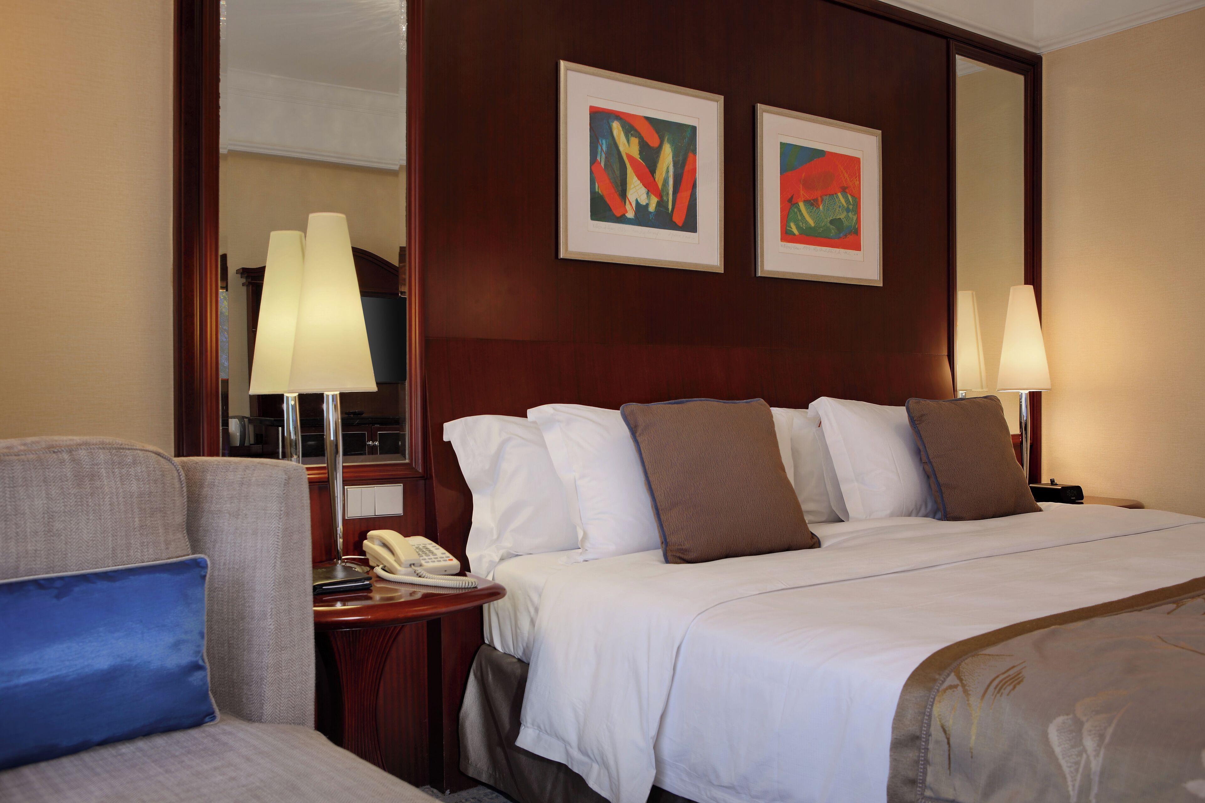 Een hotelkamer met een bed, twee bedlampen, een telefoon, een stoel en twee kunstwerken in lijsten aan de muur.