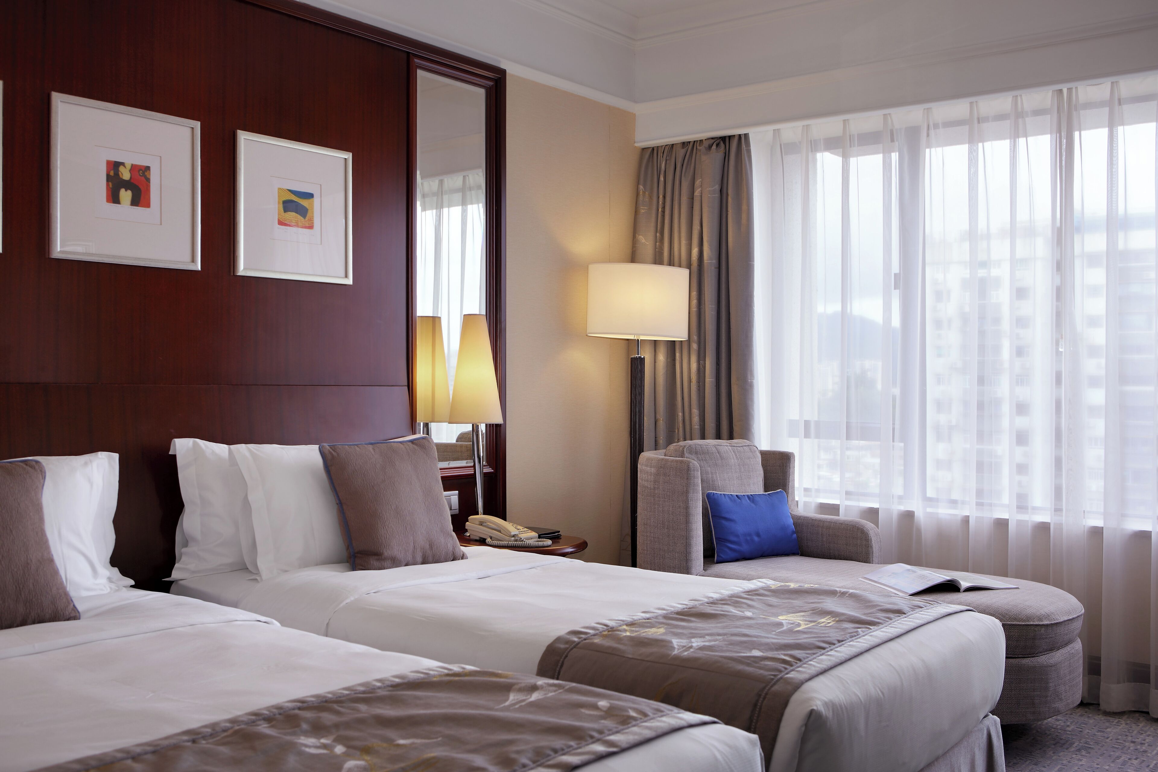 A hotel room with two beds, a sofa, a lamp, and a view of the city through large windows.