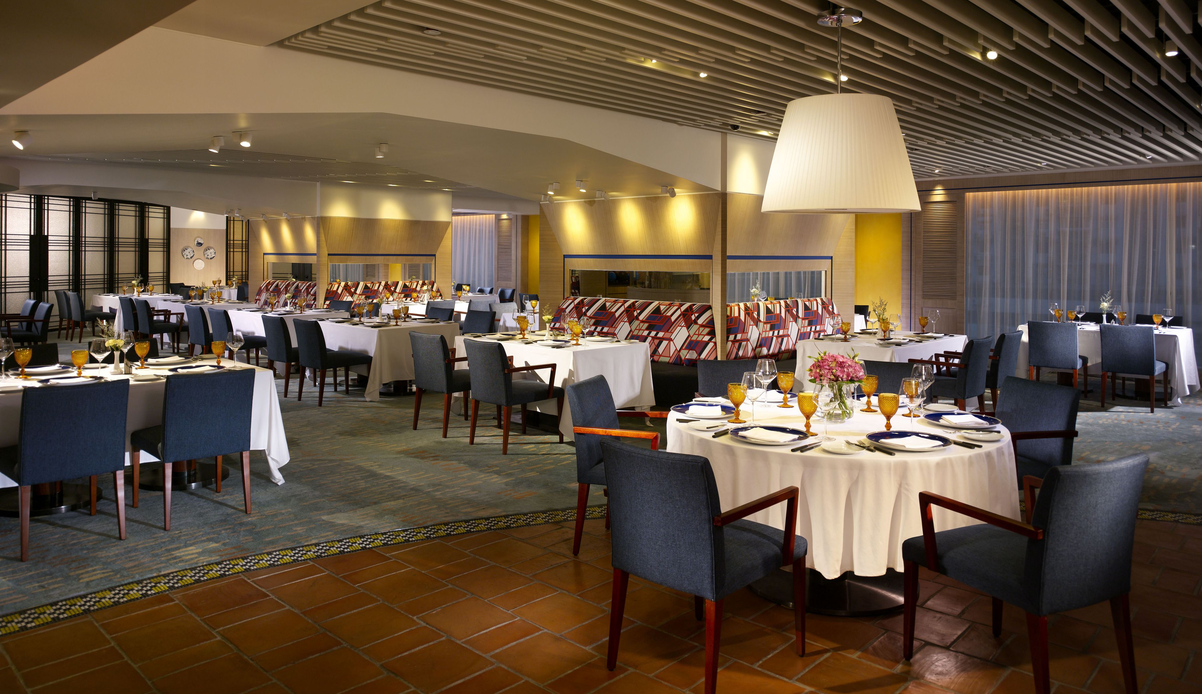 A spacious dining area with multiple tables and chairs, a large hanging light fixture, and a patterned wall design.