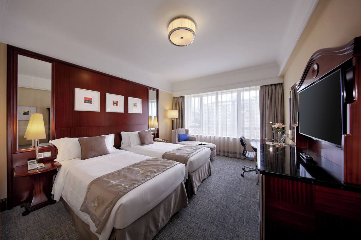 A hotel room with two beds, a TV, a desk, and a large window.