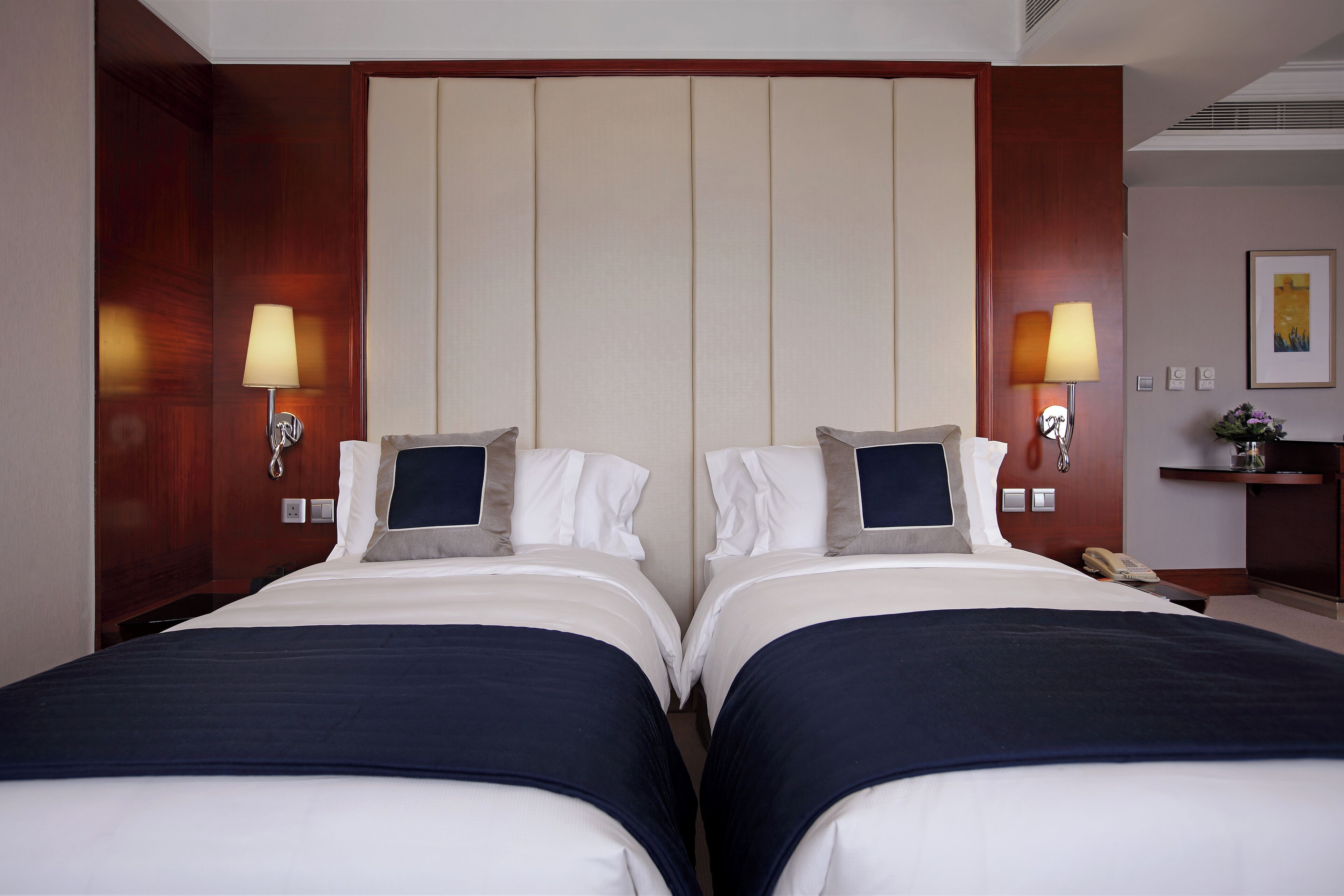 A hotel room with two beds, each with a pillow and a blanket. There are two wall-mounted lamps on either side of the beds.