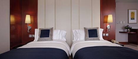 A hotel room with two beds, each with a pillow and a blanket. There are two wall-mounted lamps on either side of the beds.