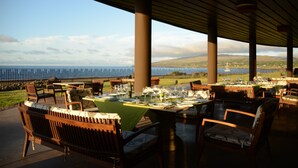 Un área de comedor al aire libre con mesas preparadas para una comida, con vista a un paisaje pintoresco del mar y las colinas.