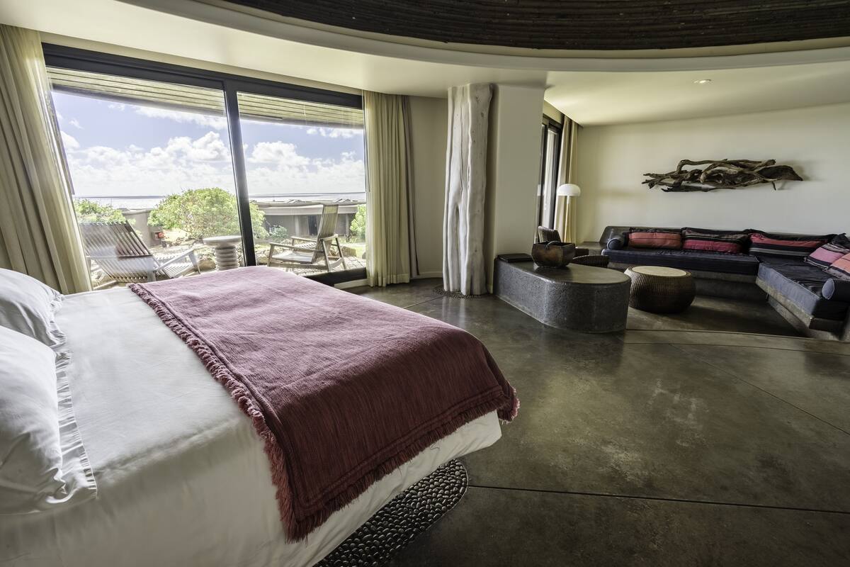 A modern hotel room with a large bed, a sofa, a coffee table, and a balcony with a view of the sky and trees.