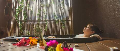 A wooden table with a plate of fruit and flowers, a bamboo screen, and a person relaxing.