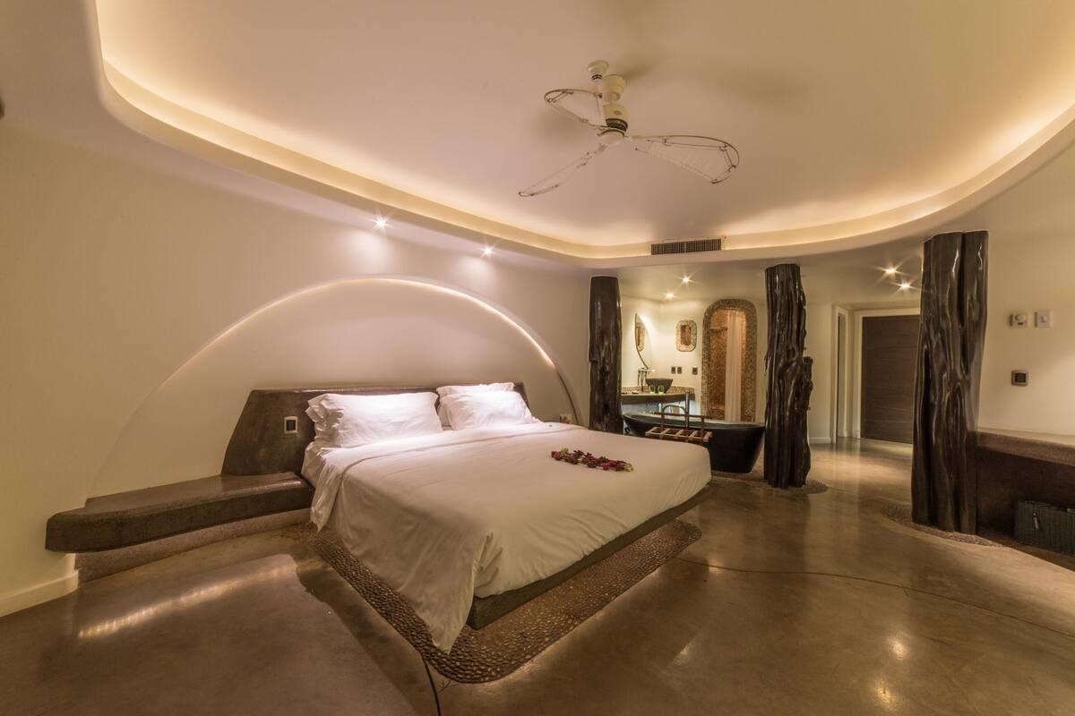 A modern bedroom with a curved ceiling, a bed with a headboard, a ceiling fan, and a bathroom with a bathtub and mirror.