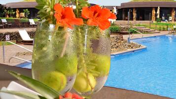 Une scène au bord de la piscine, avec une table où l’on aperçoit une boisson agrémentée de fruits et de fleurs, une assiette remplie d’un assortiment de fruits et de chocolats, et une vue sur la piscine et l’ensemble du complexe hôtelier.