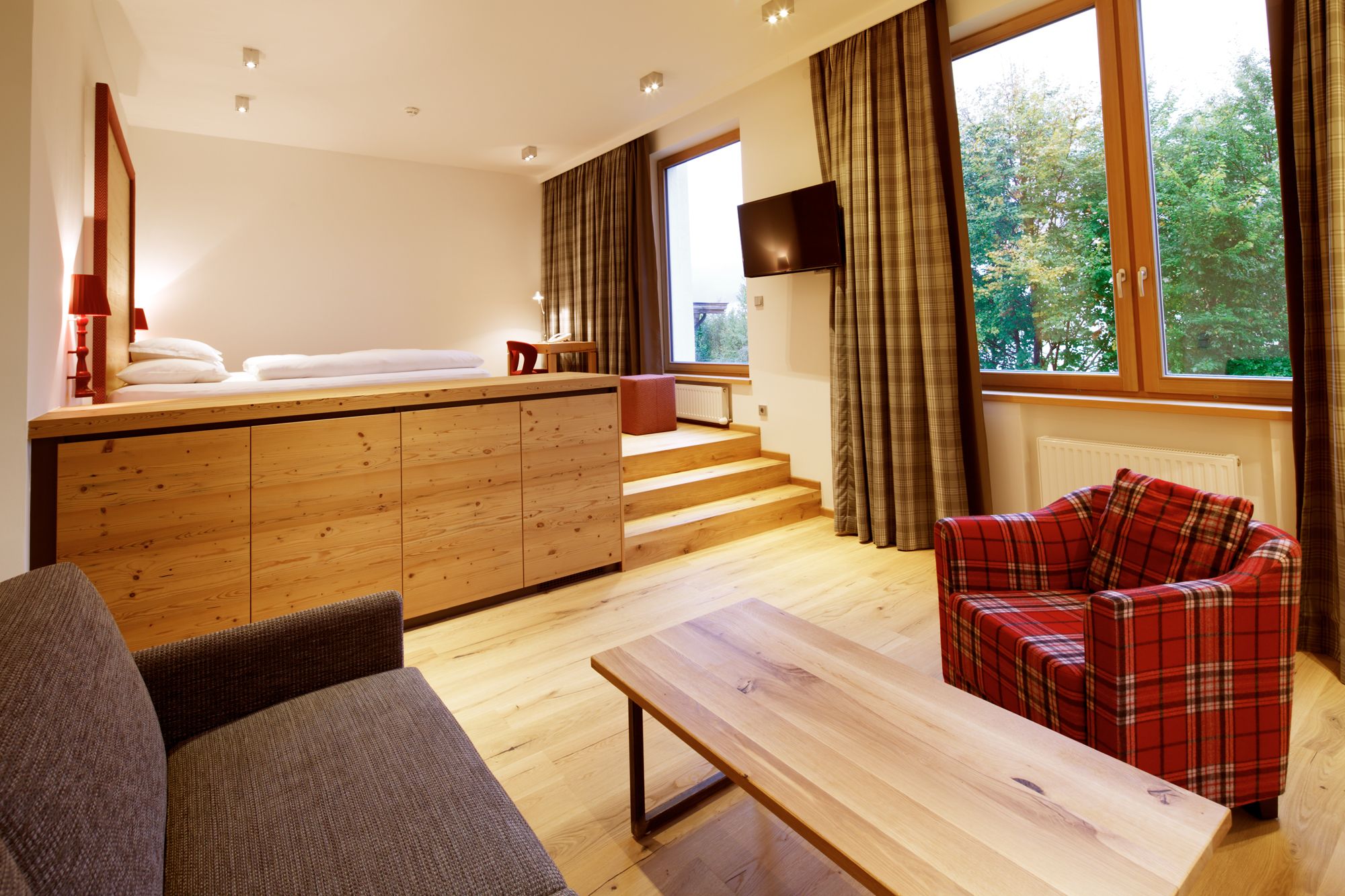 A modern hotel room with a wooden bed, a sofa, a red plaid sofa, a wooden coffee table, and a TV mounted on the wall.