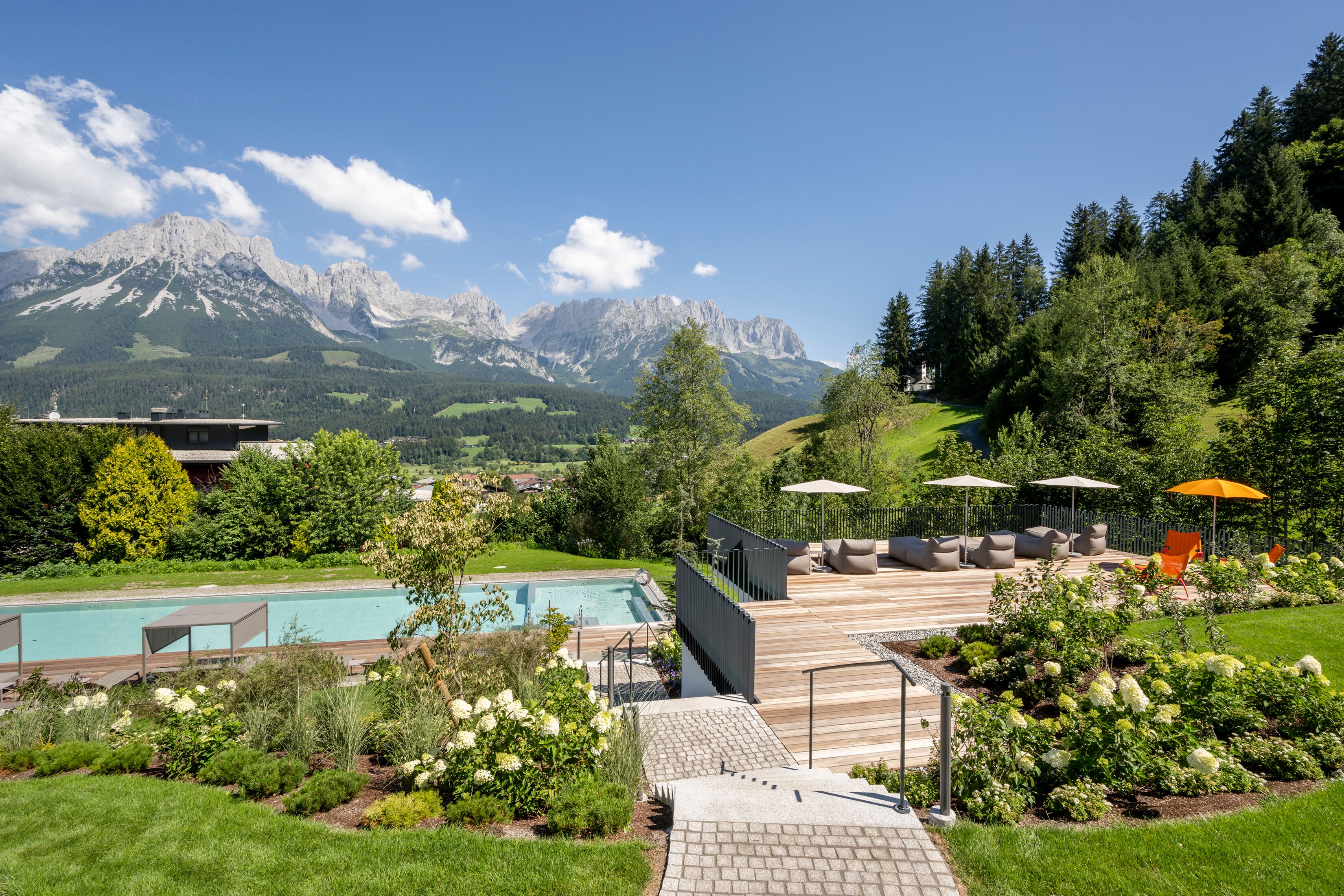 Zona de piscina con terraza de madera, mobiliario exterior y vistas a montañas.