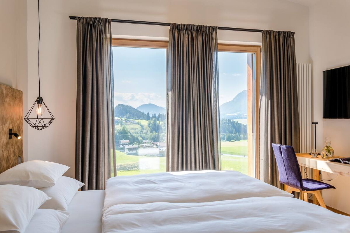 Une chambre d’hôtel avec un grand lit, un bureau et une chaise. La fenêtre offre une vue sur les montagnes et un lac.