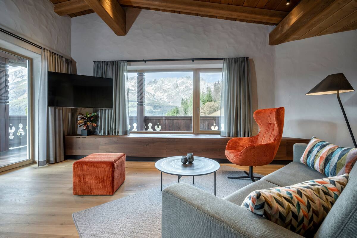 Un salon moderne doté d’une grande fenêtre, de poutres apparentes en bois au plafond, d’un téléviseur à écran plat, d’une table basse ronde et d’un canapé agrémenté de coussins à motifs.
